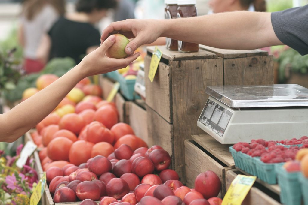 A farmer's market fruit stall vendor handing an apple to a customer.